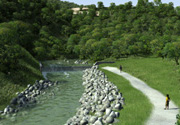 A gravel path runs alongside a flowing river with stone embankments, surrounded by lush green vegetation. Two people walk on the path under a clear sky.