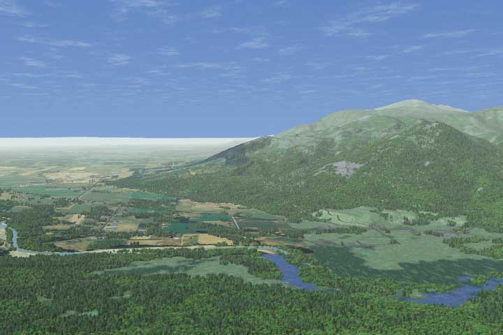 Aerial view of a landscape featuring a river, patches of forest, and fields extending towards a distant, gently sloping mountain under a blue sky with scattered clouds.